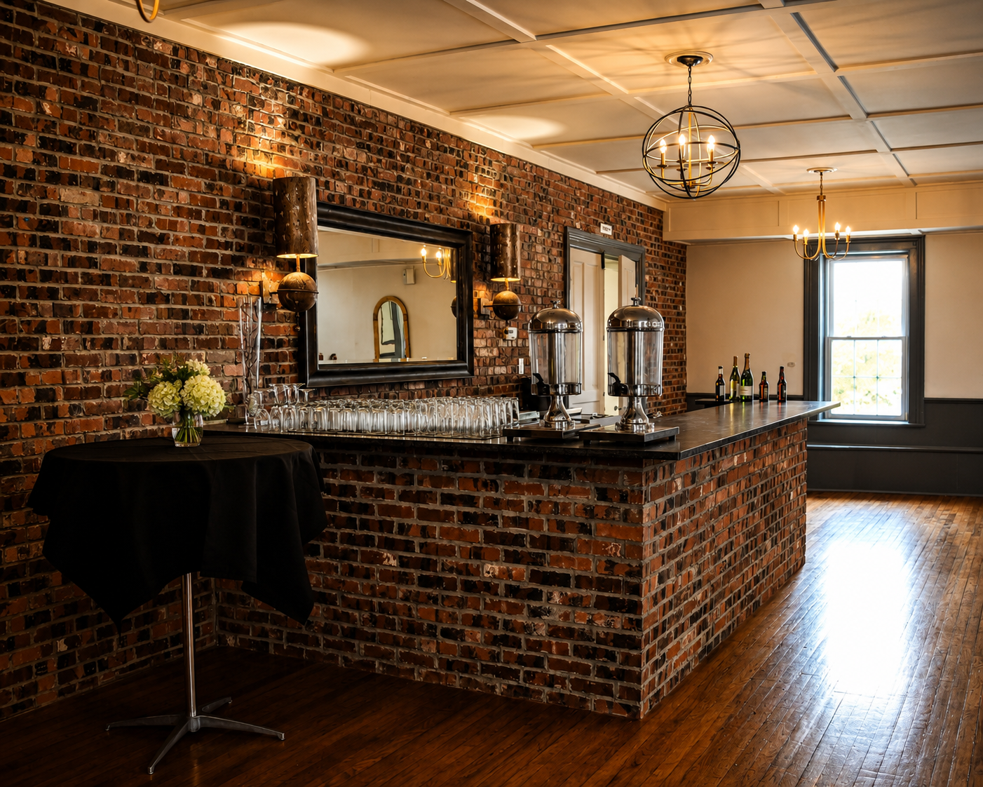 Indoor bar area with brick walls, wooden floors, chandeliers, and a large window. Bar counter with glass dispensers and bottles. Small round table with black tablecloth and flower arrangement.