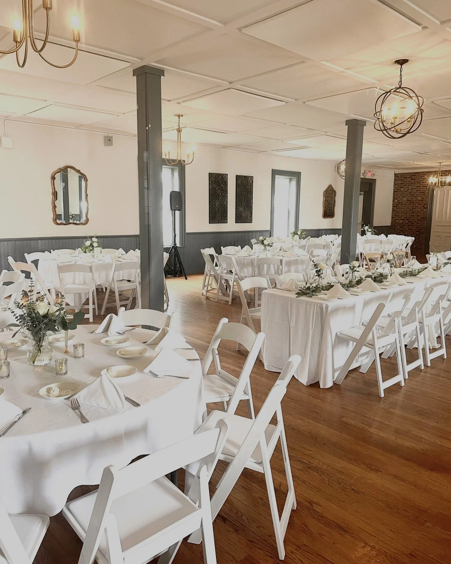 Room set up for a formal event with round tables covered in white tablecloths, decorated with floral centerpieces and candles, and white folding chairs. The room has white walls, wood flooring, and is lit with chandeliers.