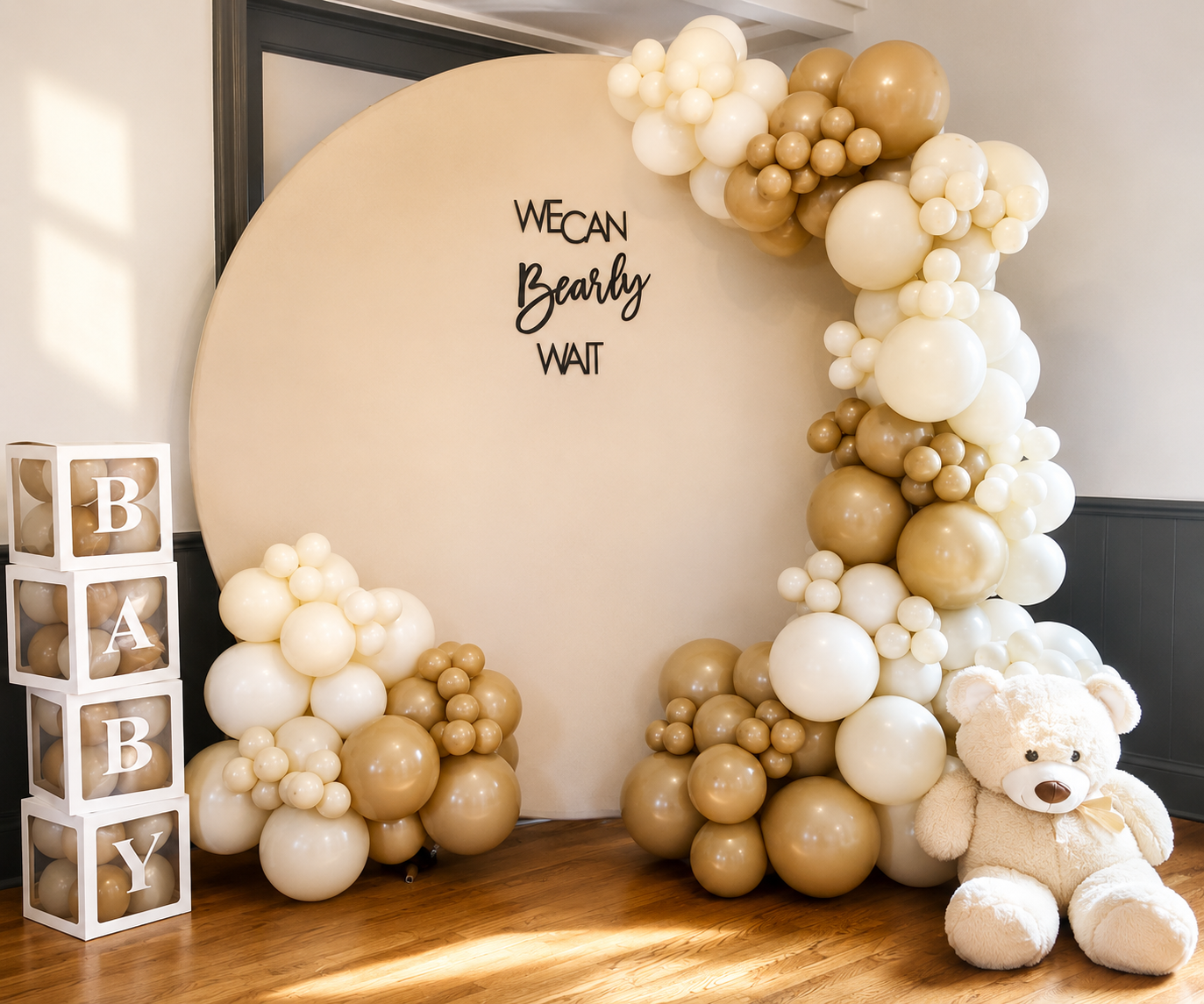 Decorative baby shower backdrop with a large white circular panel, surrounded by cream, white, and gold balloons. A teddy bear sits on the floor in front of the balloons. To the left, three cube blocks spell 'BABY' with balloons inside. The panel has