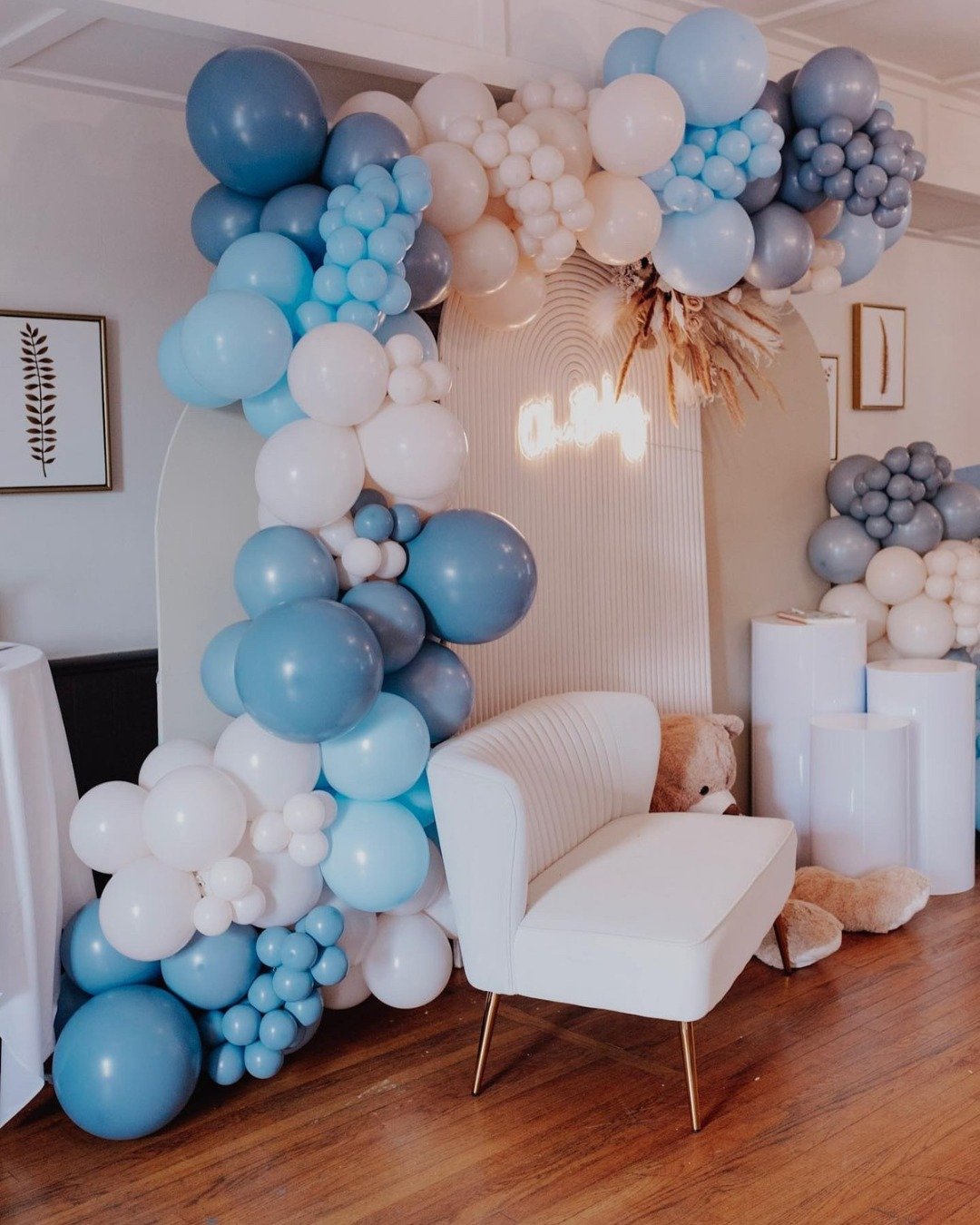 Decorative balloon arrangement in shades of blue, white, and gray with a neon sign that reads 'baby', a white upholstered bench, stuffed teddy bear, and framed images on the wall.