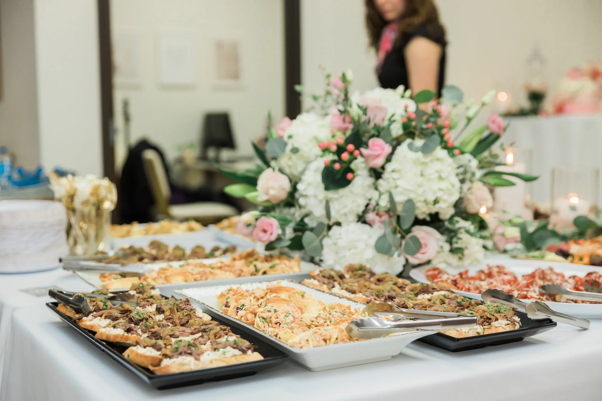 Buffet table with various dishes and a floral centerpiece in the background at an indoor event.