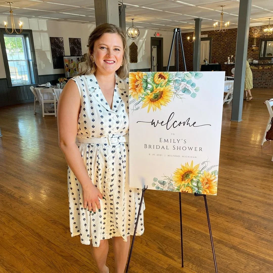 A woman smiling and standing next to a decorative welcome sign for Emily's Bridal Shower in a spacious, well-lit event hall with wooden floors and tables set for a celebration.