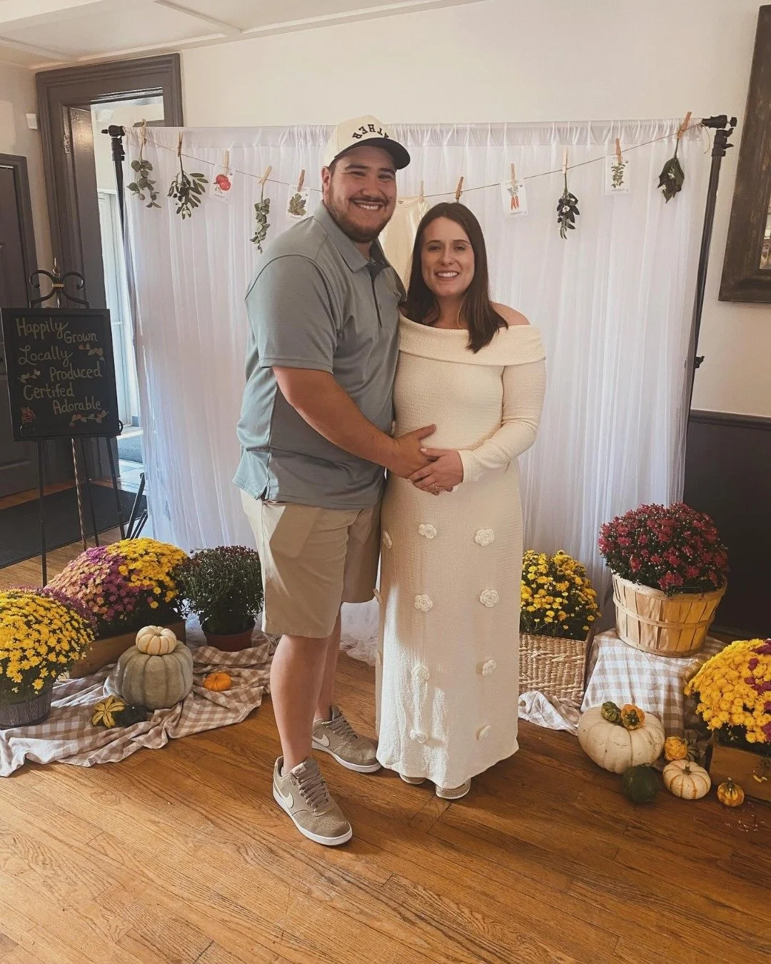 A man and woman smiling and holding hands in front of a white curtain backdrop decorated with hanging leaves and cards, surrounded by pumpkins and potted chrysanthemums on the floor.