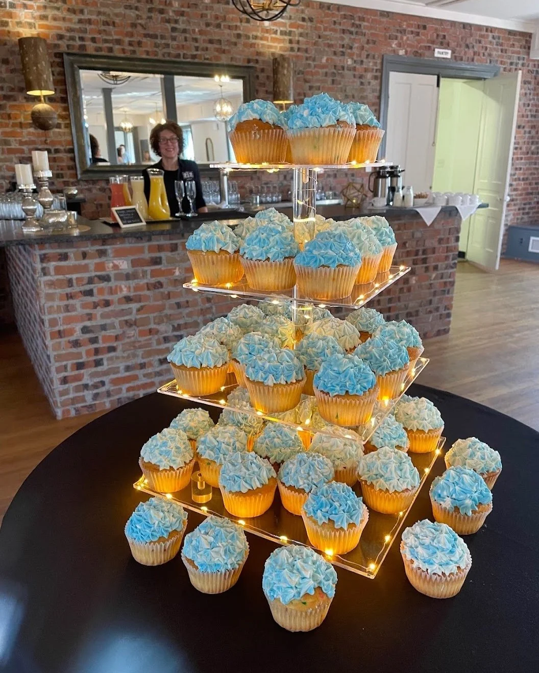 Cupcakes with blue and white frosting arranged on a three-tiered illuminated stand at a celebration event in a room with brick walls and a bar area.