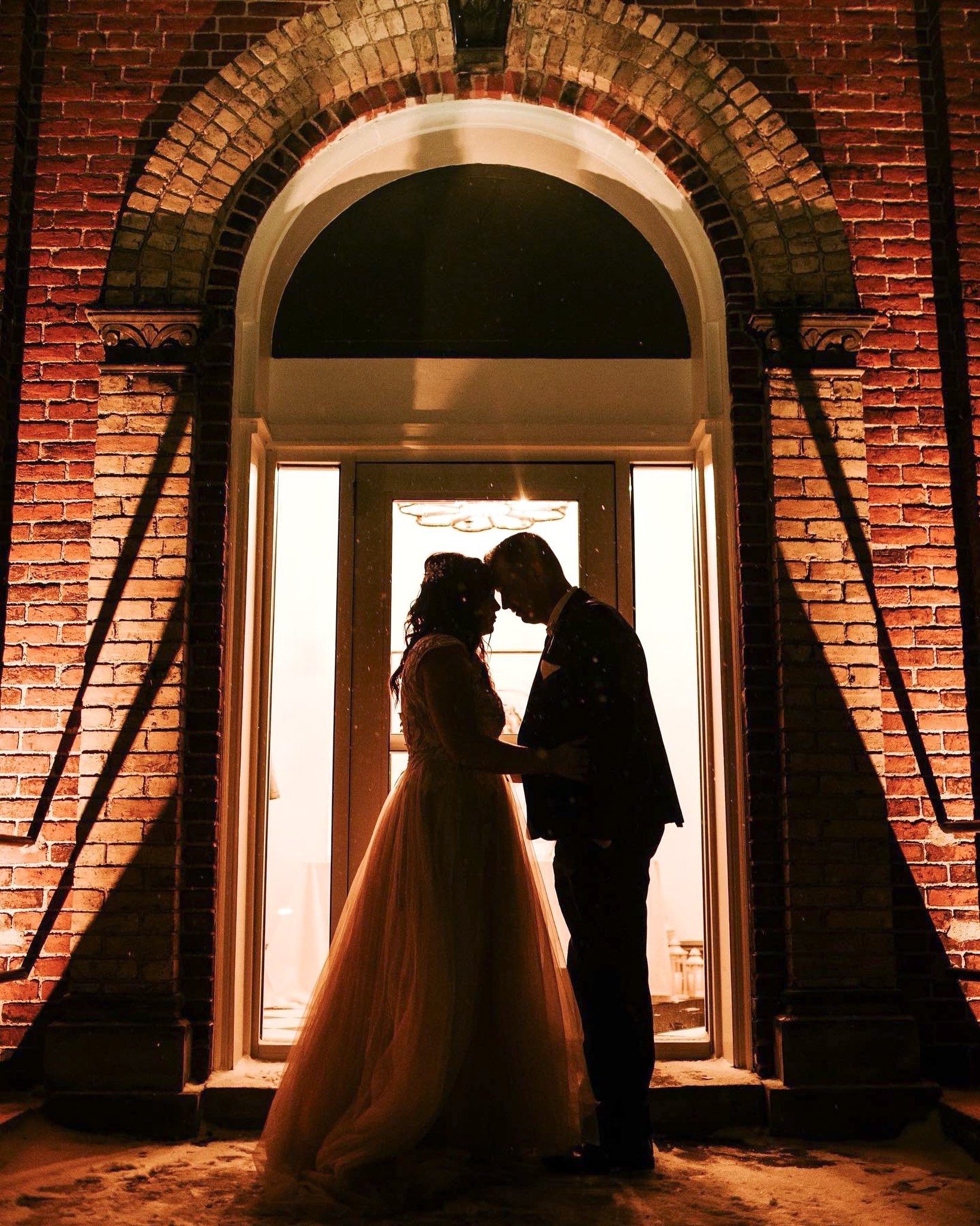 A couple stands in front of a large window at night, their foreheads touching and hands clasped. Their silhouettes are visible against the bright window light, and they appear to be sharing an intimate moment.