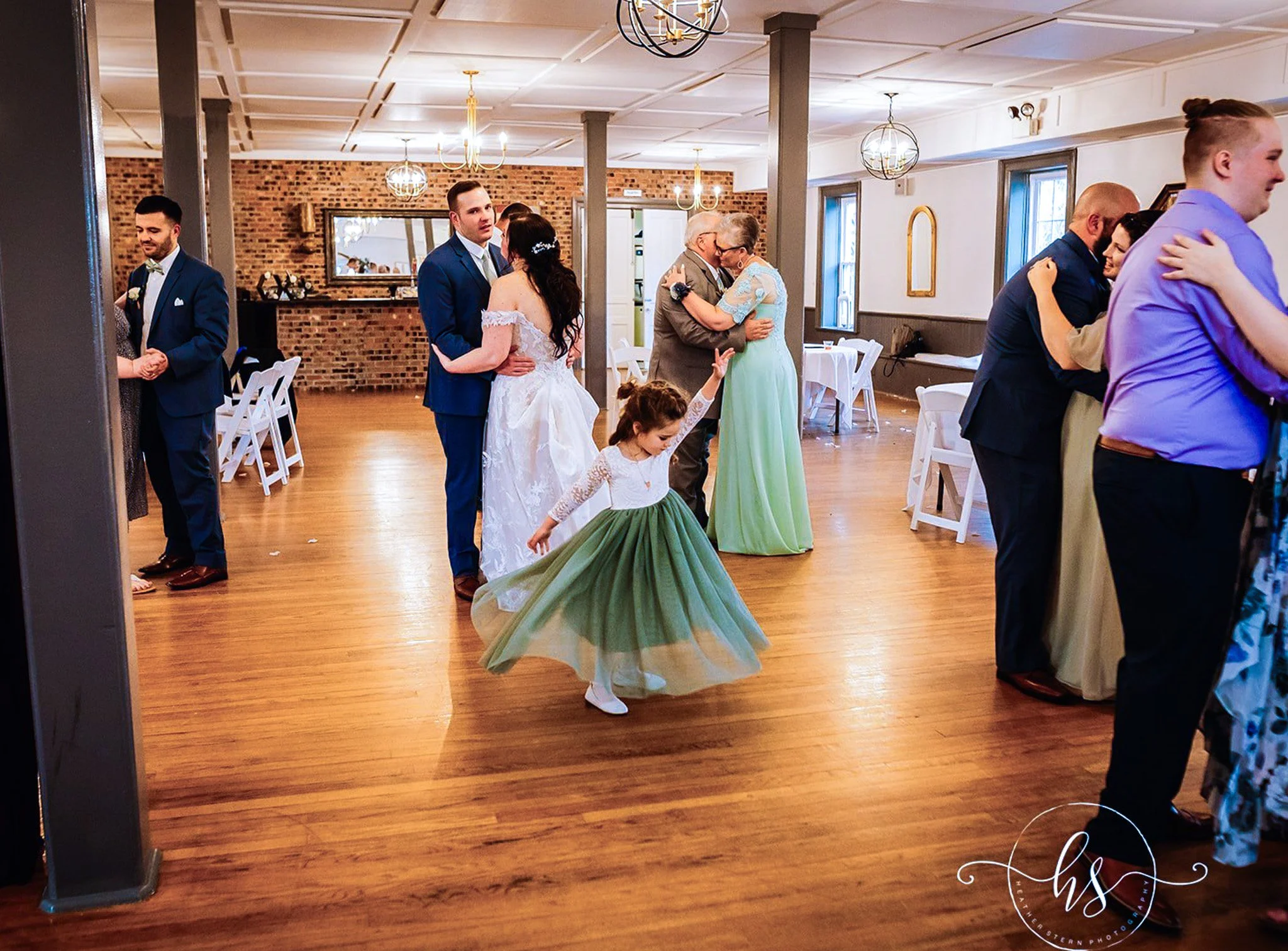 People dancing at a wedding reception in a rustic venue with brick walls, wooden floors, and soft lighting from chandeliers. The focus is on a woman in a green dress spinning with her arm raised, surrounded by couples dancing.