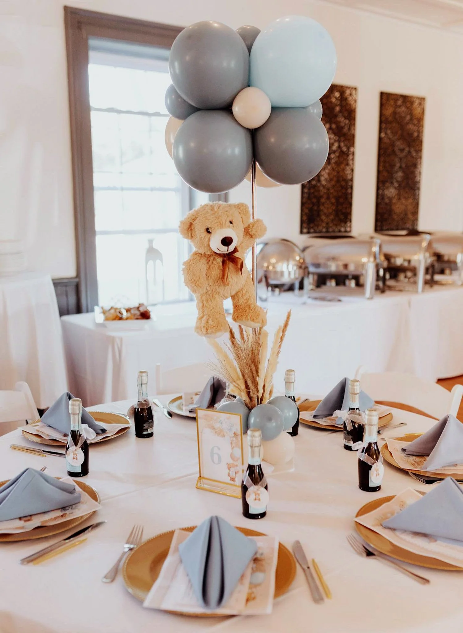 Table decorated with a teddy bear, gray and white balloons, champagne bottles, and place settings for a celebration or party.
