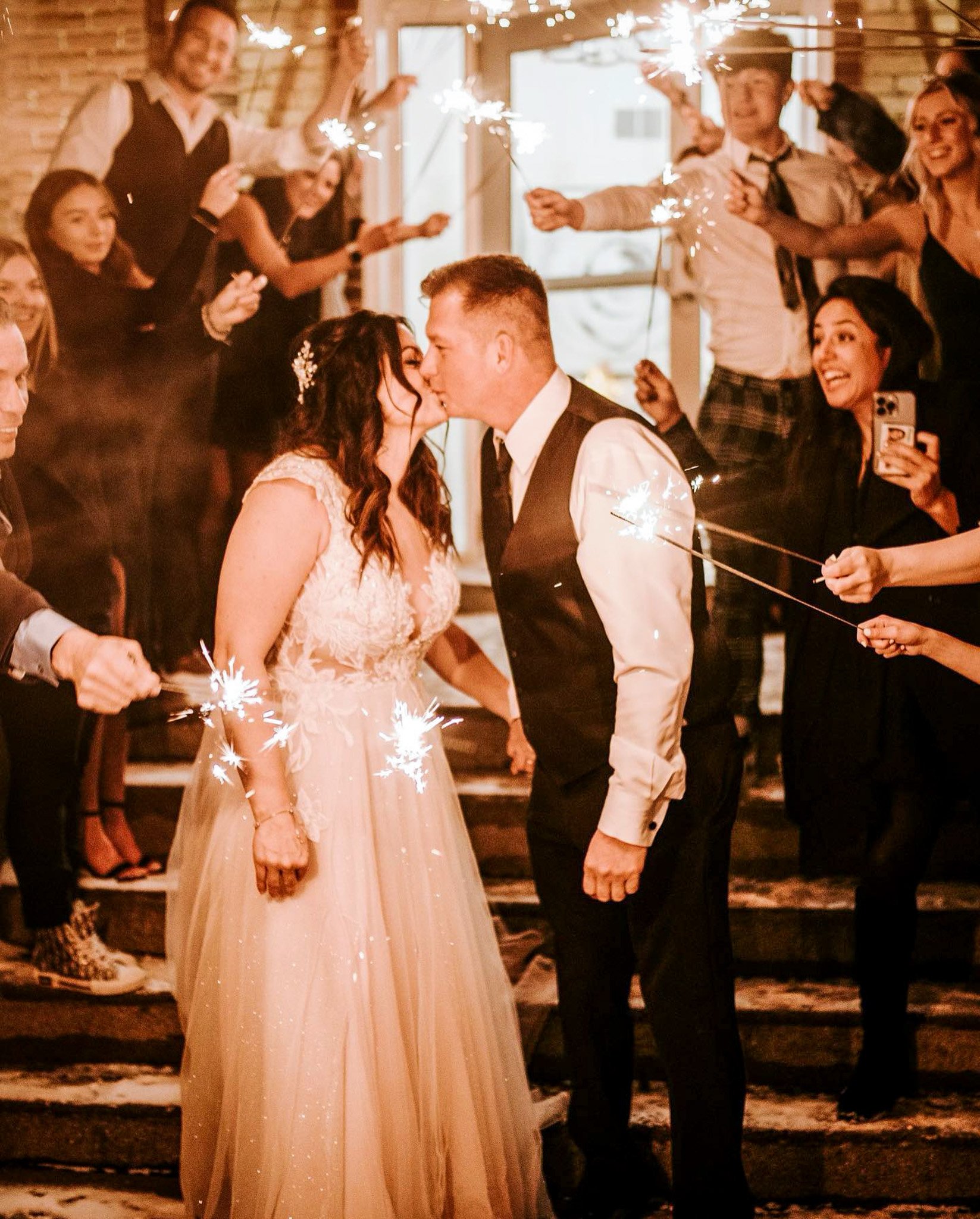 A bride and groom kiss at their wedding, surrounded by friends holding sparklers and celebrating indoors with warm lighting and wooden walls.