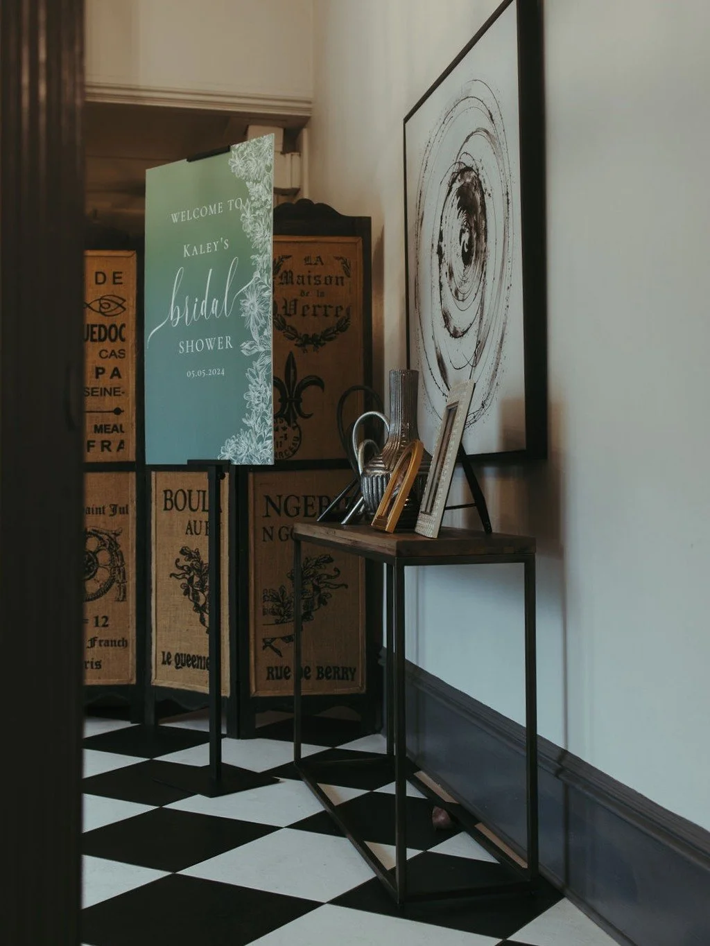 Wedding shower welcome sign in a hallway with black and white checkered floor, behind a decorative room divider, with framed art and metal vases on a small table.