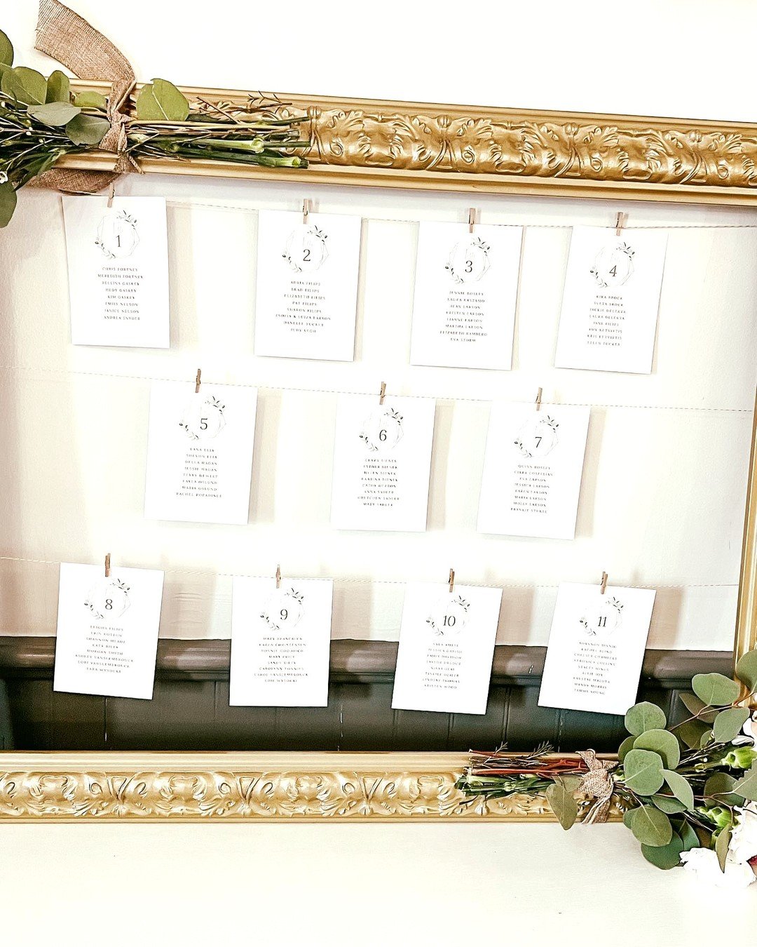 Wedding seating chart with numbered cards hanging on strings, decorated with greenery and floral arrangements, framed with an ornate gold frame.
