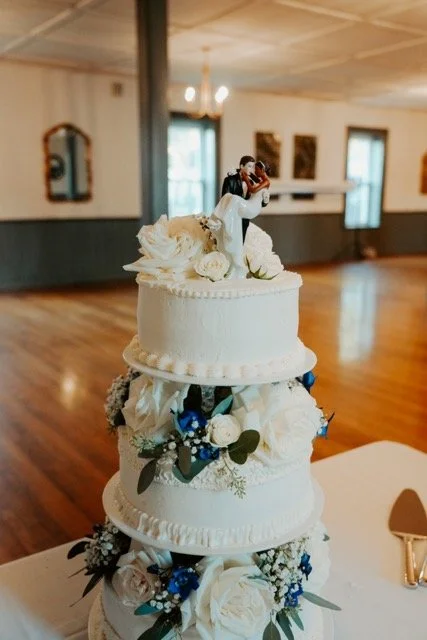 Three-tier white wedding cake decorated with white and blue flowers and a bride and groom cake topper, set in a reception hall with wooden floors and large windows.