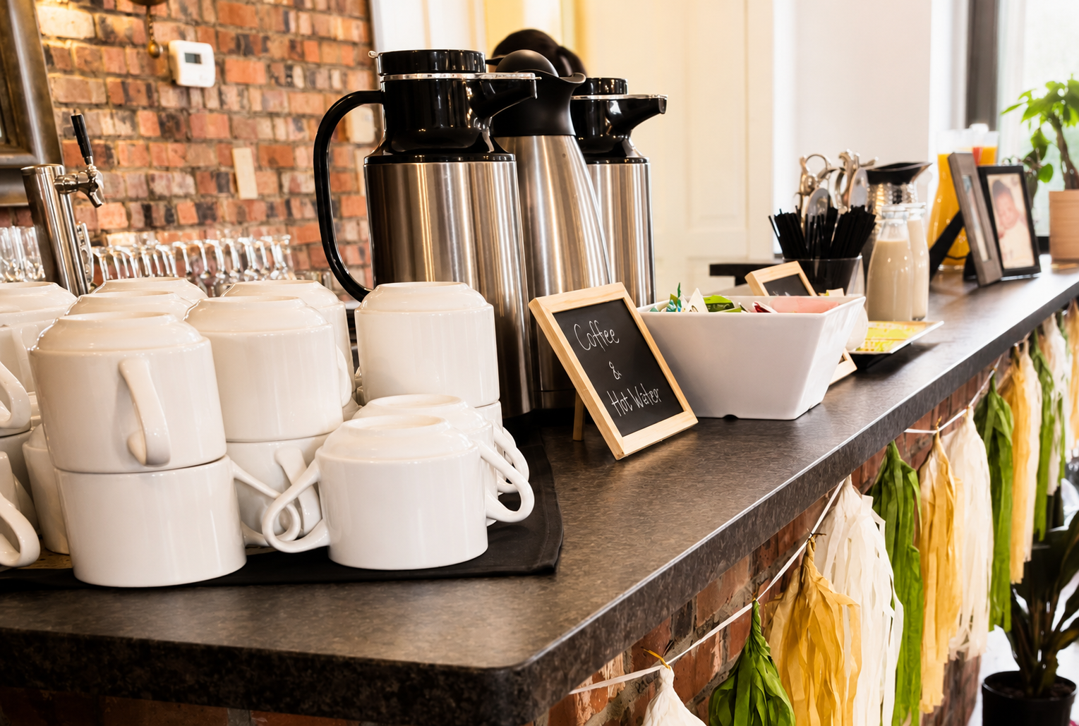 Coffee station with white mugs, stainless steel hot water dispensers, a bowl of condiments, and a small chalkboard sign reading "Coffee & Hot Water" in a cafe with a brick wall background and decorative garland underneath the counter.