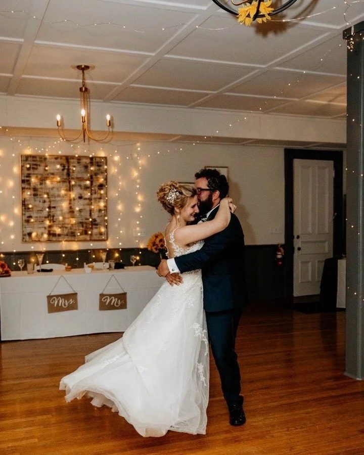 A bride and groom dance together, smiling at each other in a decorated reception hall with string lights and a draped table in the background.