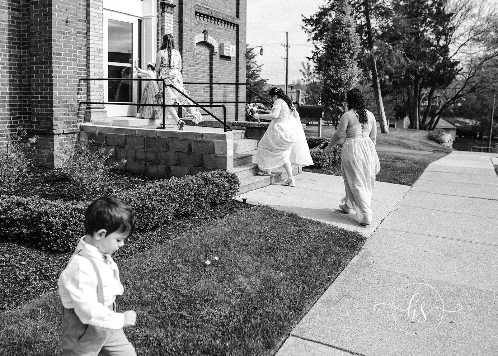 People in wedding attire walking up steps to a brick building with a glass door, a young child in the foreground on grass, and trees in the background.