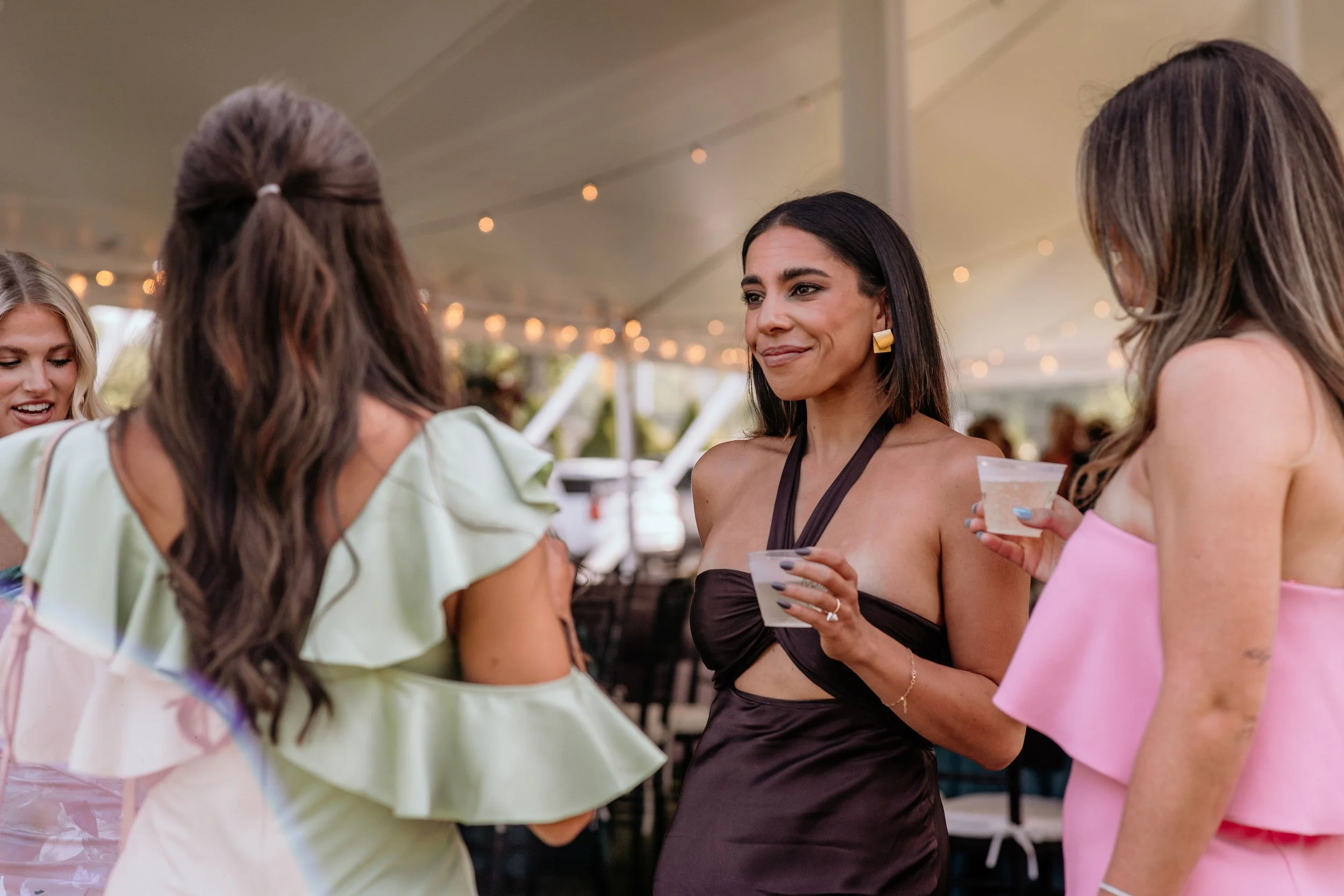 A group of diverse women at an outdoor event, socializing under string lights, holding drinks and engaging in conversation.