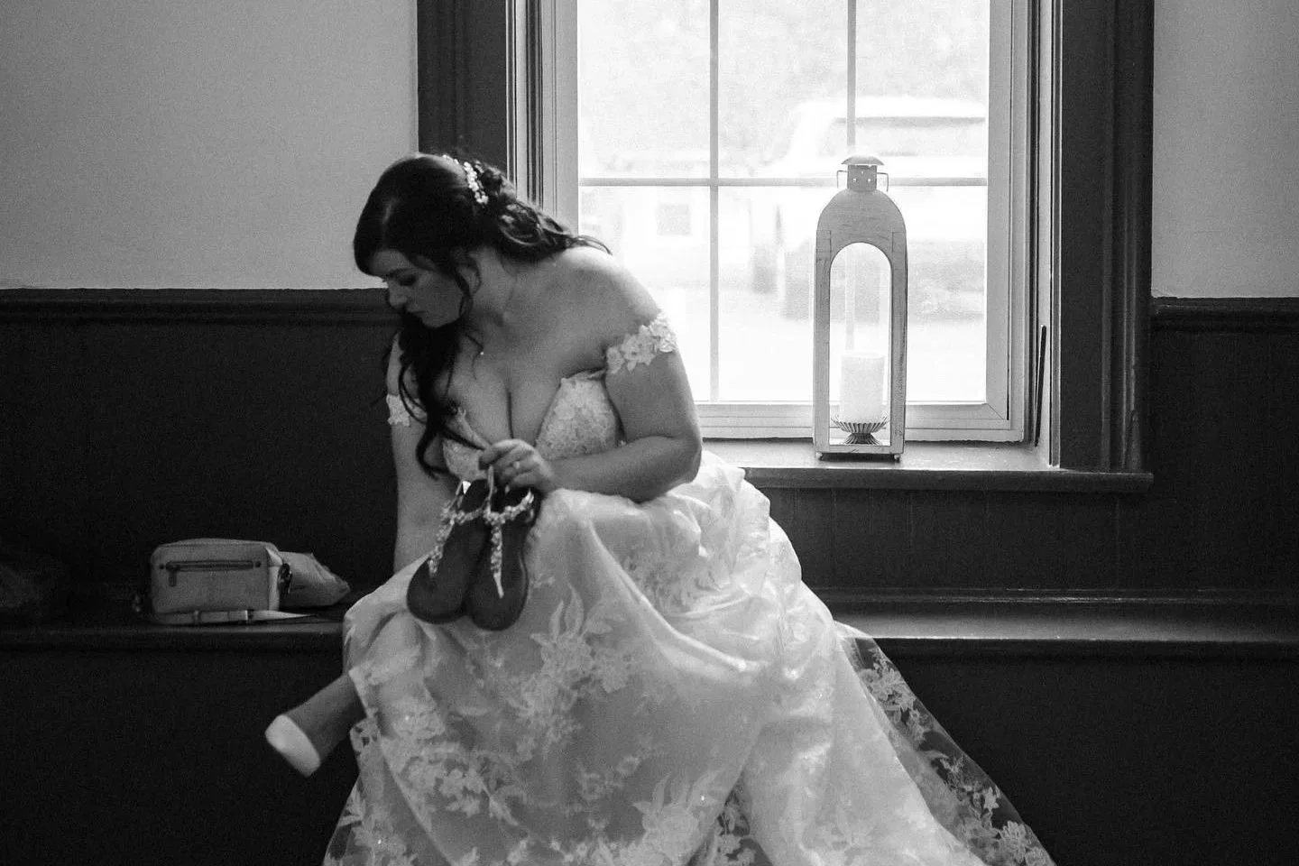 A woman in a wedding dress sitting on a window sill, holding wedding shoes, with a lantern on the window ledge in black and white.