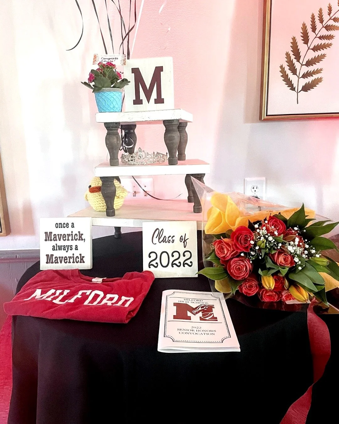 Senior graduation display with a black table covered with a red and black cloth, featuring a red Milford hoodie, a graduation program, and signs reading 'once a Maverick always a Maverick' and 'Class of 2022'. A bouquet of red roses and yellow tulips
