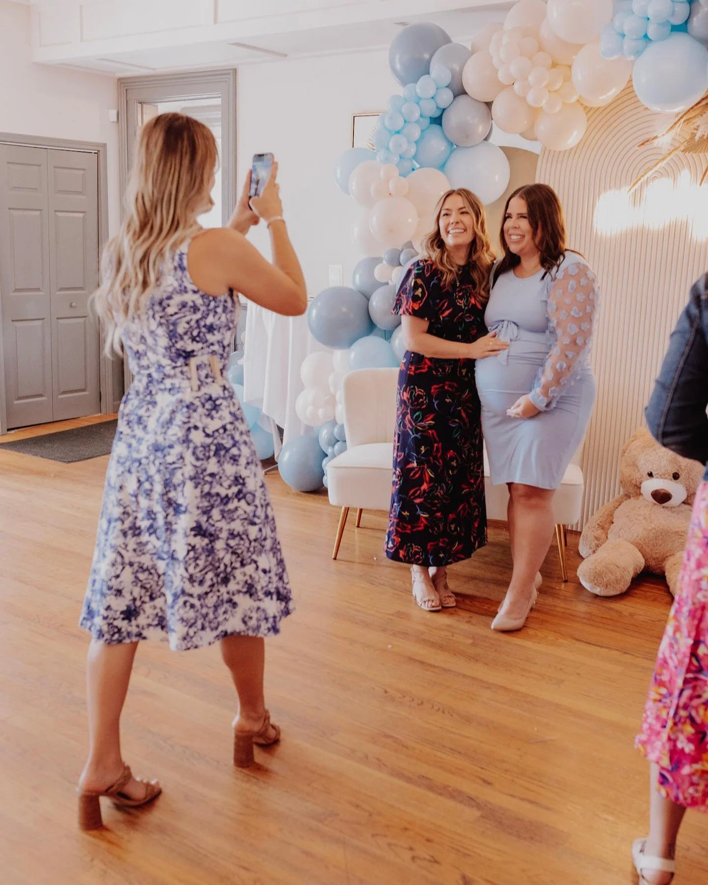 Two women posing for a photo at a celebration with balloon decorations, one woman is wearing a floral dress and the other is wearing a light blue dress with sheer sleeves with floral patches, another woman in a floral dress is taking their picture, a