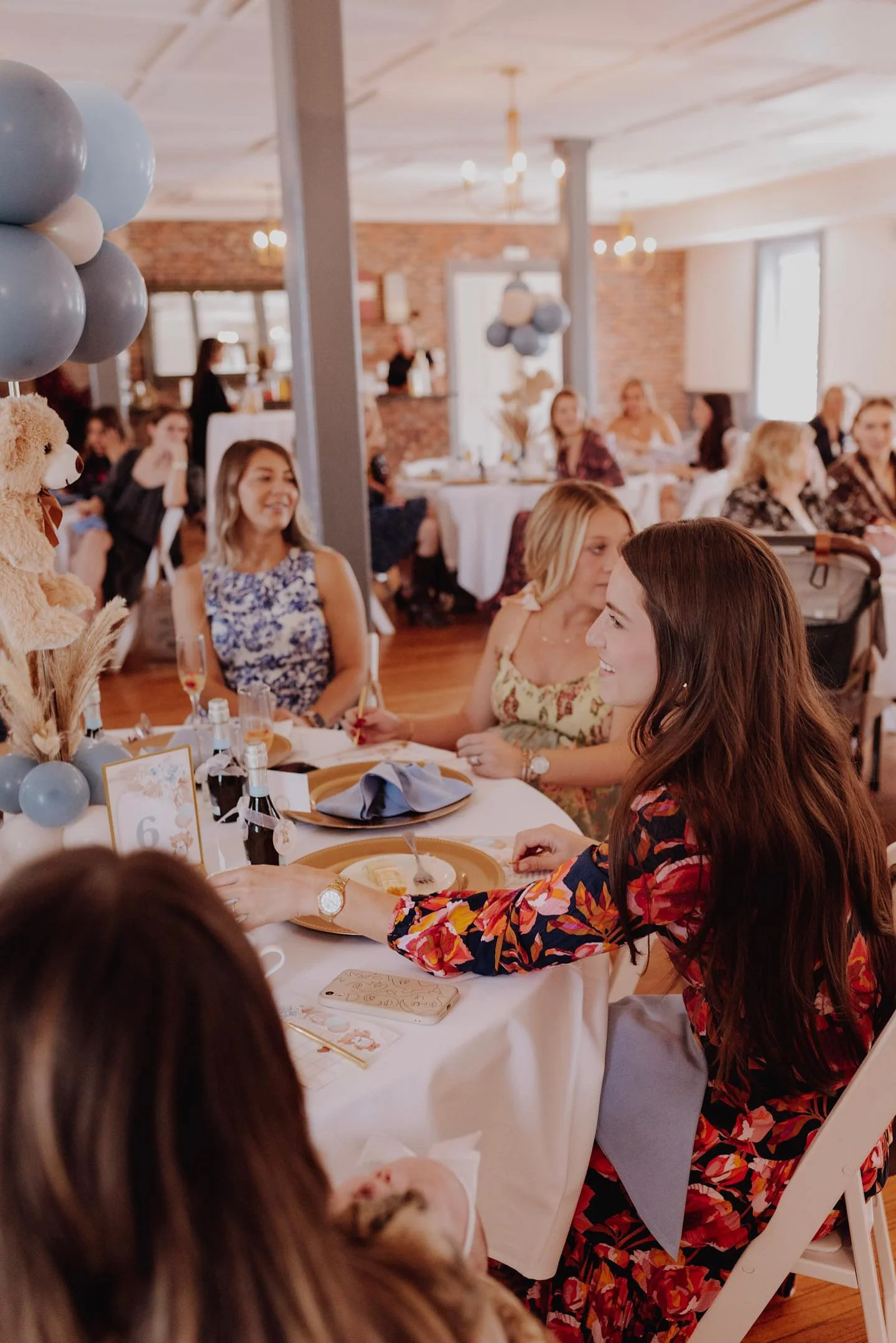 A gathering of people at a decorated indoor party with balloons and floral arrangements, enjoying food and socializing.