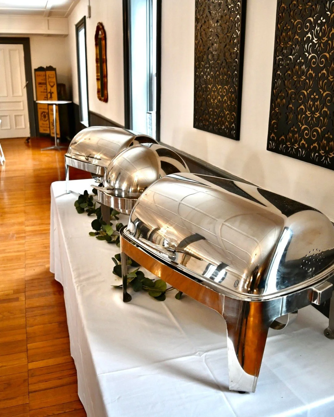 Three stainless steel chafing dishes on a white tablecloth, decorated with greenery, in a room with wooden floors, windows, and decorative wall art.