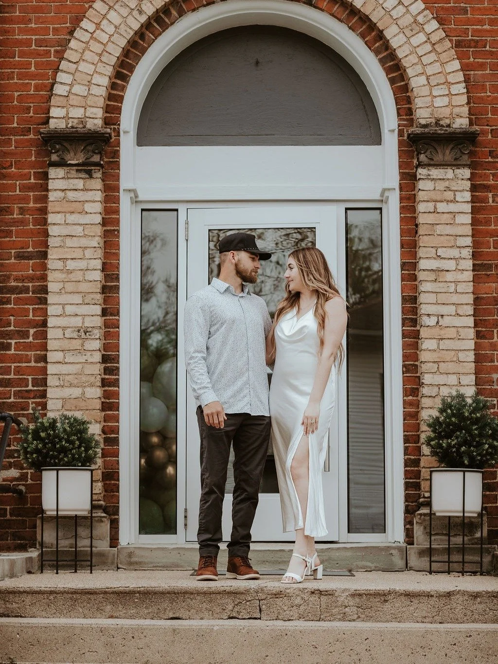 A couple standing outside a brick building with a glass door, looking at each other. The woman wears a white dress with a slit, and the man wears a light-colored shirt with dark pants and a cap. Potted plants are on either side of the door.