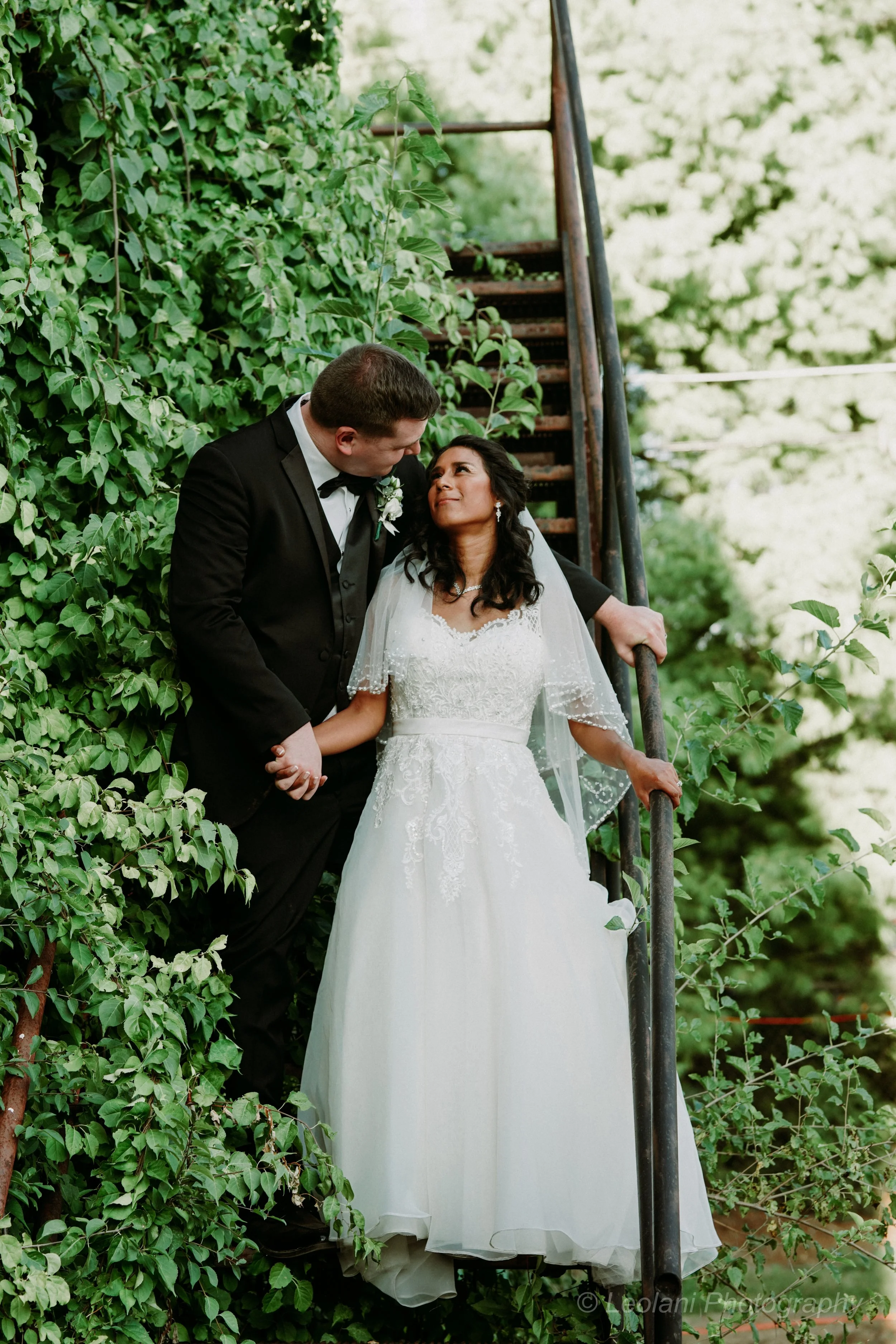 A wedding couple stands on a fire escape, surrounded by greenery, with the groom in a black tuxedo and the bride in a white wedding dress, holding hands and looking at each other.