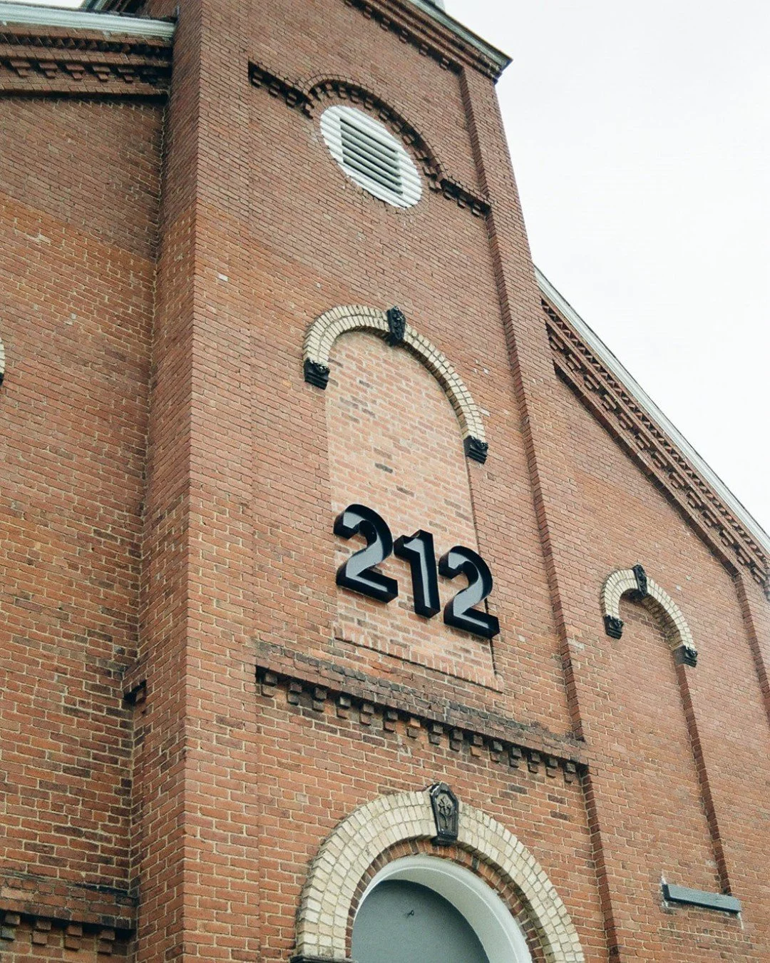 Close-up view of a brick building with the number 212 in bold black letters on it. The building has arched windows and decorative brickwork.