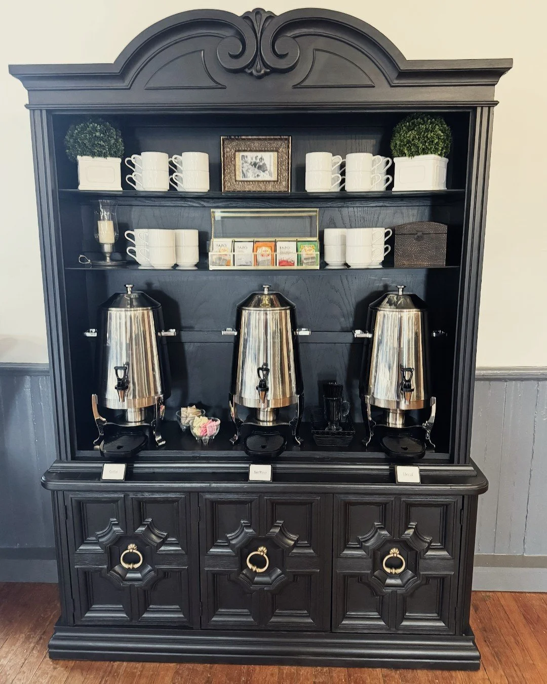 A black wooden coffee station with three stainless steel coffee dispensers, cups, and tea boxes on shelves.
