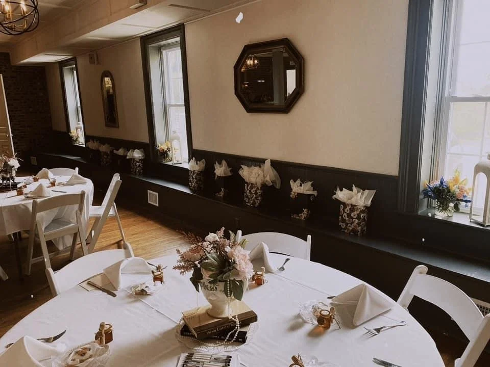 Interior of a well-decorated dining room with round tables, white tablecloths, folded napkins, and floral centerpieces, with natural light coming through the windows.