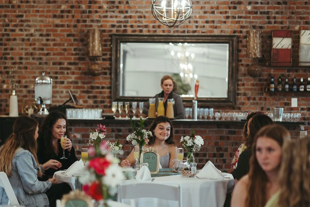 A group of women sitting at a decorated table in a restaurant with a brick wall background, and a woman in a gray shirt standing behind a bar in the background.