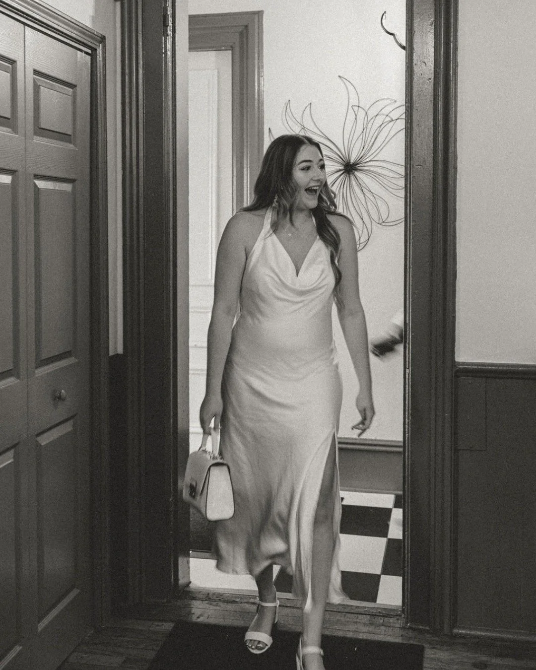 A woman in a satin dress with a high slit is smiling and walking through a doorway, carrying a small handbag and wearing heels.