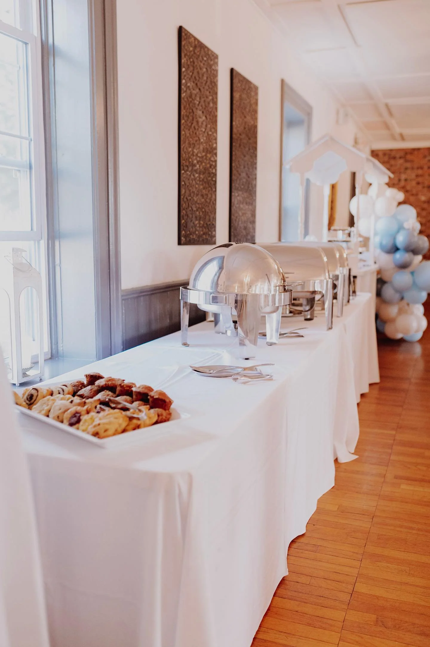 Buffet table with cookies, chafing dishes, and balloons in the background at a celebration event.
