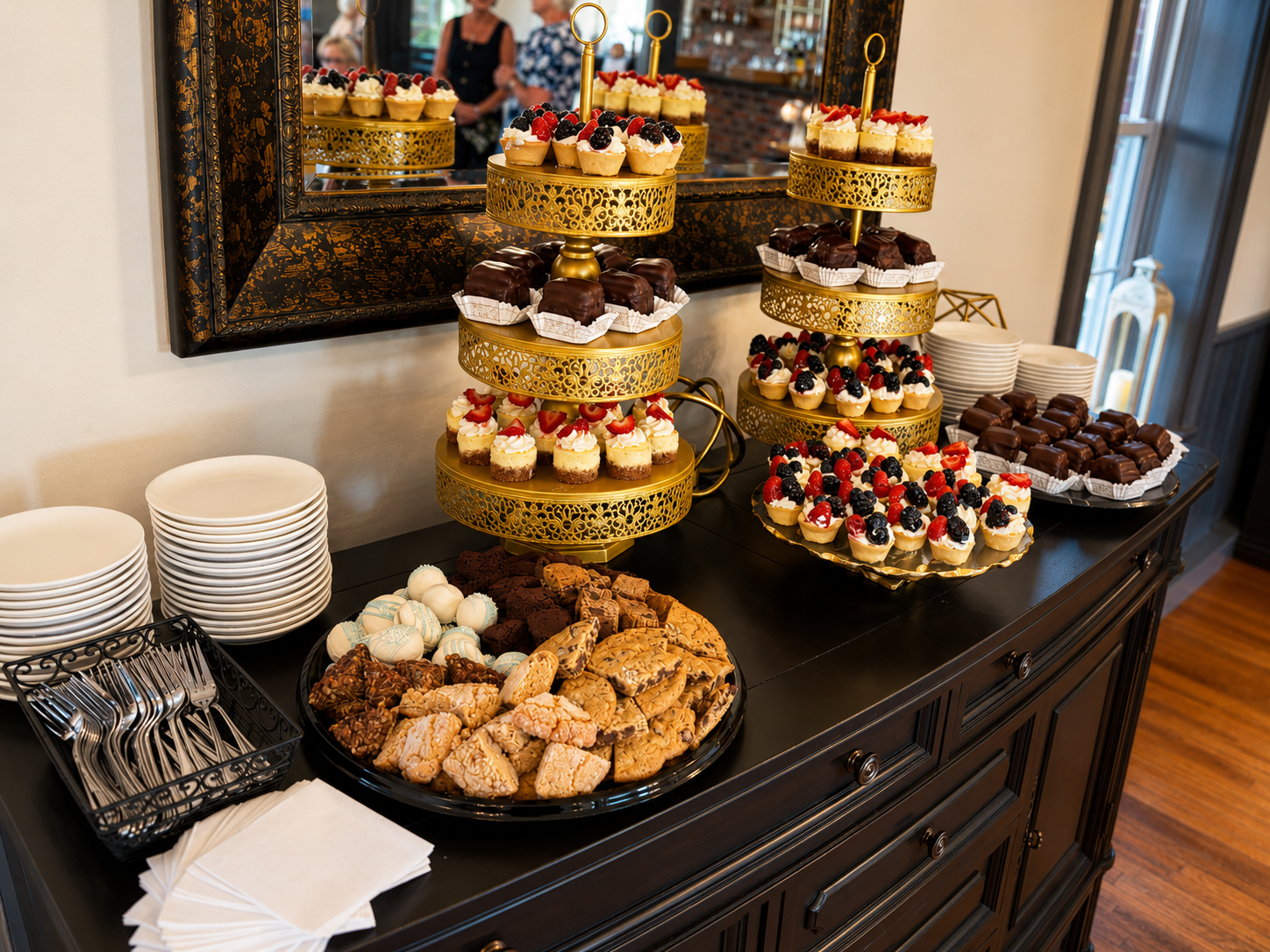Dessert table with assorted pastries, including fruit-topped mini cheesecakes, chocolate-covered cakes, and cookies, arranged on gold tiered trays and black plates. Stacked white plates, silverware, and napkins are on the side. A mirror behind reflec