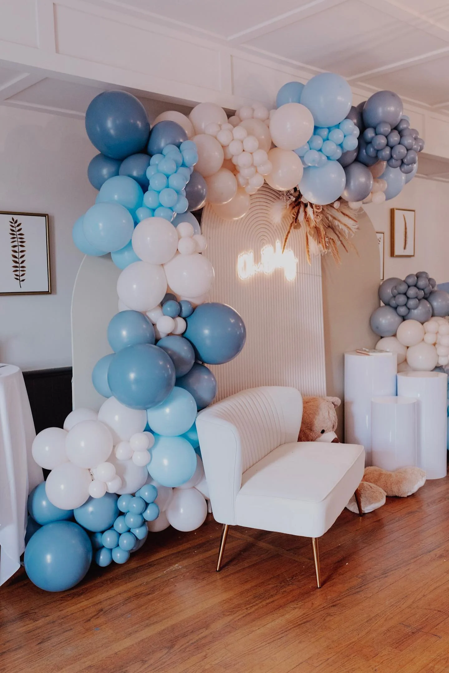 A balloon arch with white, light blue, gray, and darker blue balloons decorates a room with white walls and a ceiling. There is a white upholstered chair with metal legs in front of the balloon arch. The room has wooden flooring, framed artwork on the walls, and a plush teddy bear on the floor next to the chair.