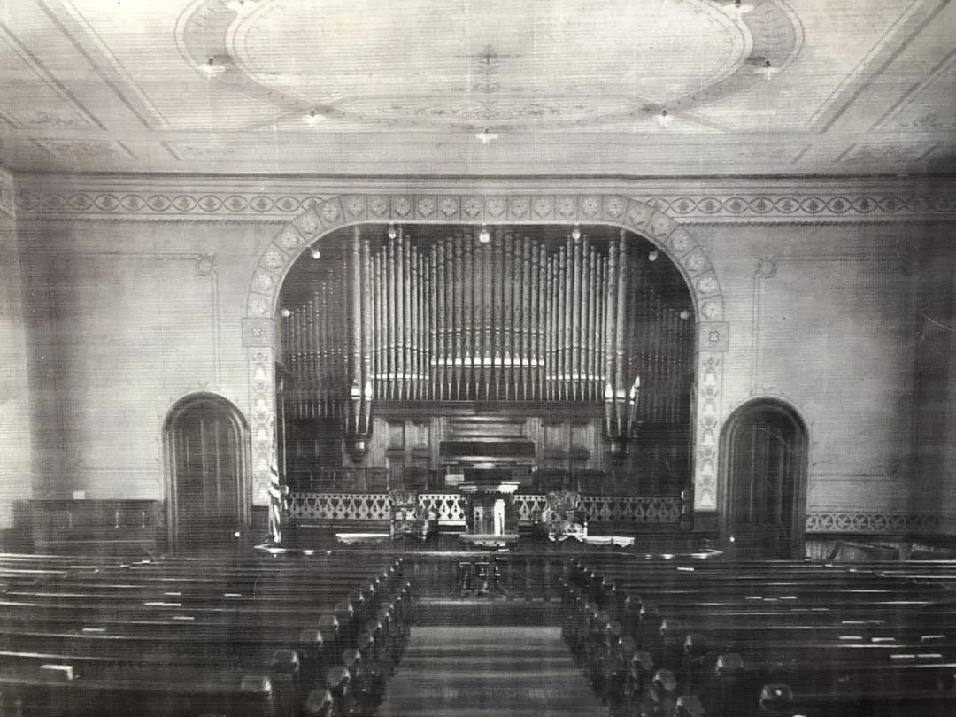 An empty church sanctuary with rows of wooden pews facing a stage with a large pipe organ.