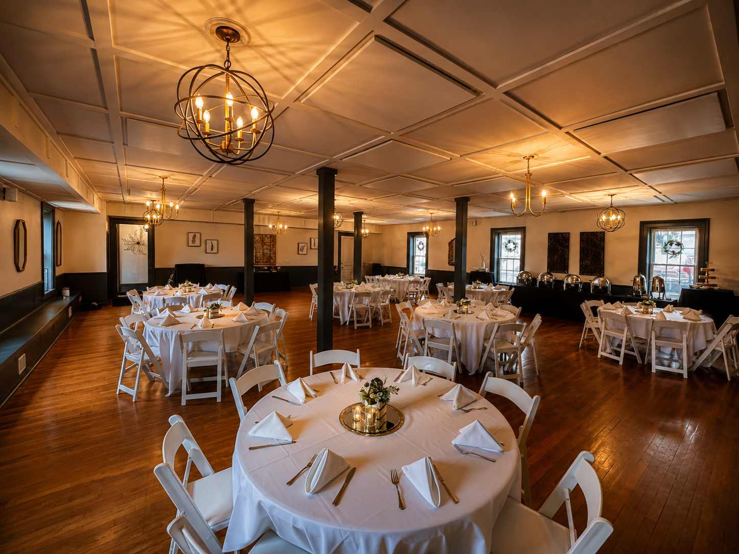 A banquet hall set up for an event with round tables covered in white tablecloths, each with folded napkins, cutlery, and a small floral centerpiece. The room has wooden floors, black and white walls, multiple windows with holiday wreaths, and chande
