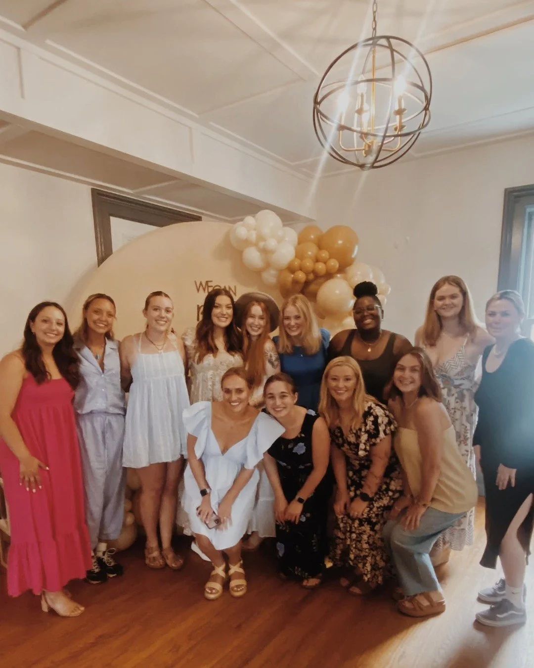 Group of diverse women posing indoors at a celebration with balloon decorations behind them.