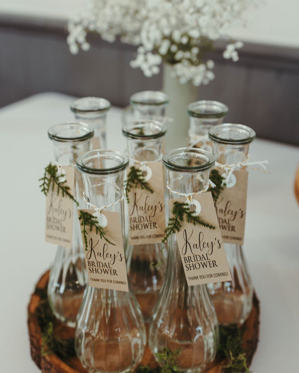 Nine empty glass bottles with labels reading "Kale's Bridal Shower" and tiny green foliage, arranged on a wooden base, with white flowers in the background.