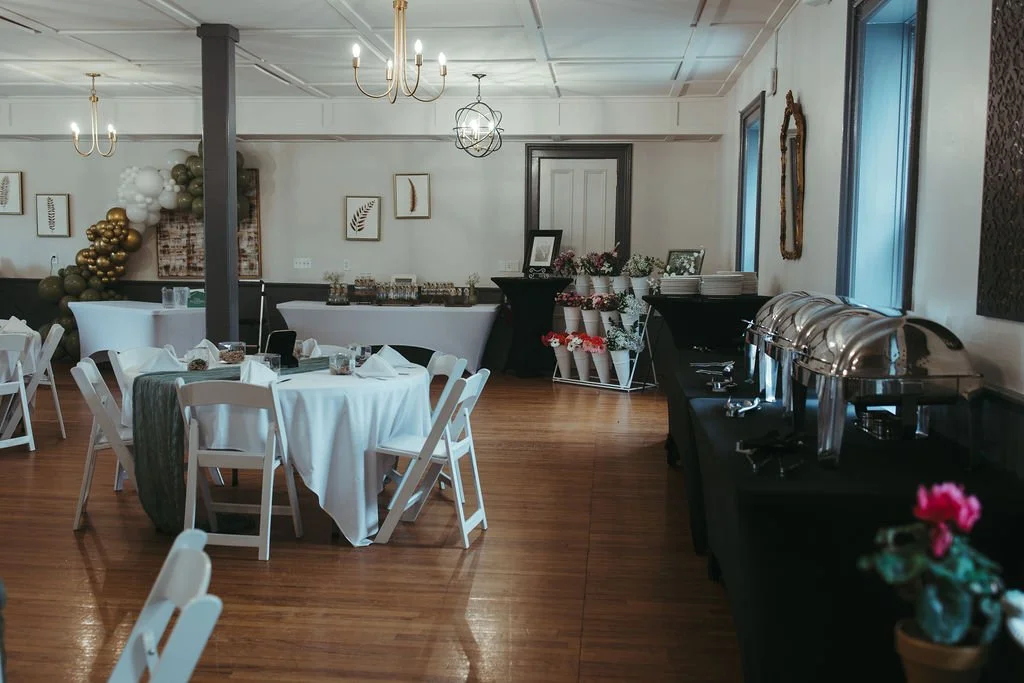 Event space with decorated tables, a balloon arrangement, and a buffet setup on the side. Wooden floors and framed artwork on the walls.