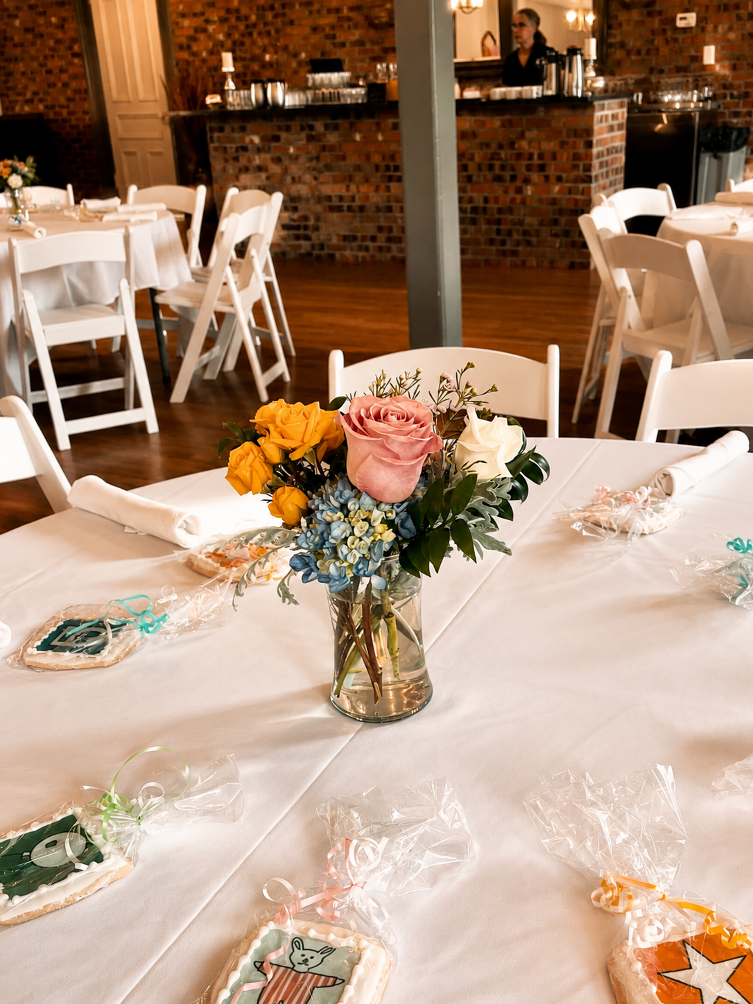 A decorated event table with a bouquet of pink, yellow, and white roses, and other flowers in a glass vase. The table has small decorated cookies wrapped in plastic with ribbons. In the background, there are additional tables with white chairs and a 
