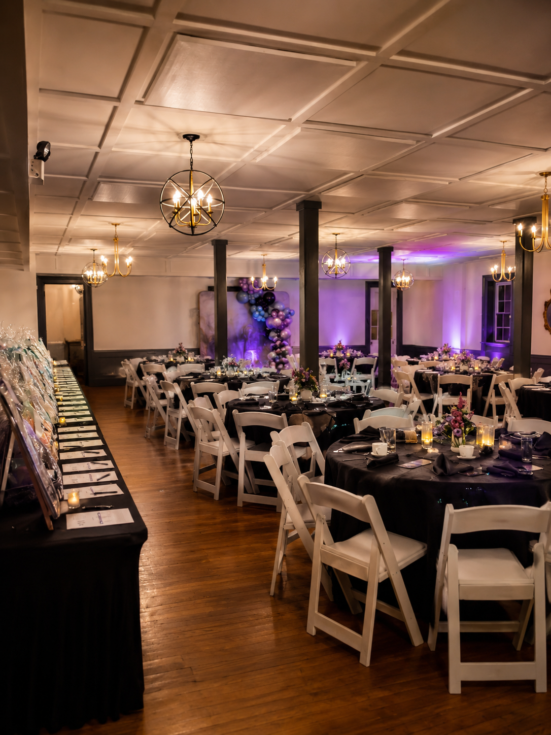 Elegant banquet hall decorated for an event with round tables covered in black tablecloths, white chairs, floral centerpieces, and candles. The room has a wooden floor, chandeliers, and purple lighting on the walls, with balloons arranged on a backdr