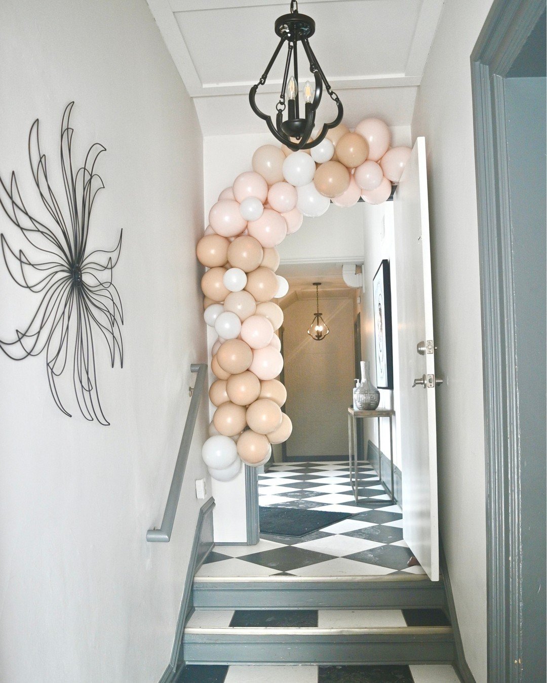 Decorated hallway with balloon arch in shades of beige, white, and blush pink, a black chandelier, a wall art piece resembling a butterfly, and a black and white checkered floor.