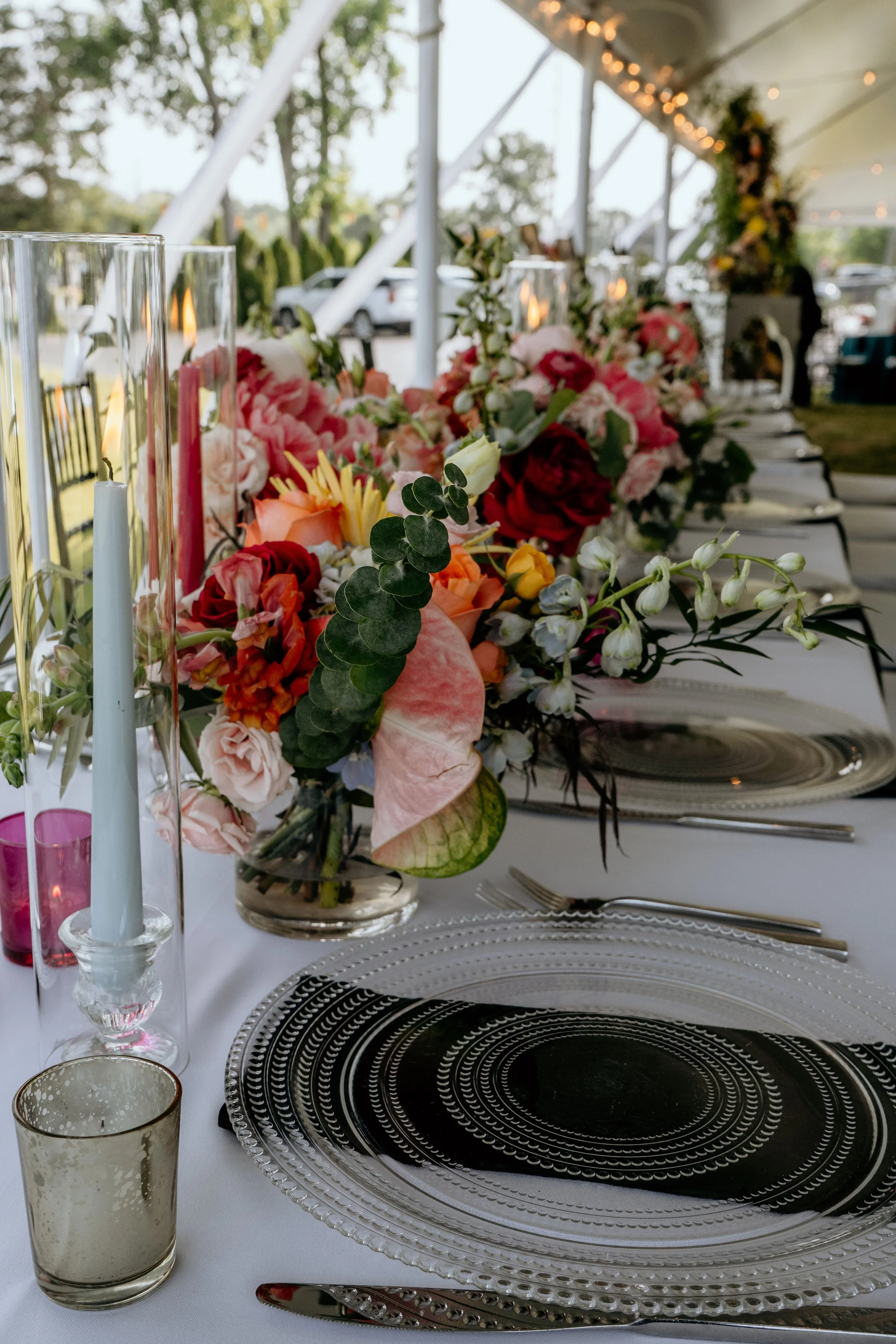 A close-up of a table setting at an outdoor event, featuring a floral centerpiece with pink and red flowers, candles in glass holders, silverware, and decorative plates with black and silver designs.