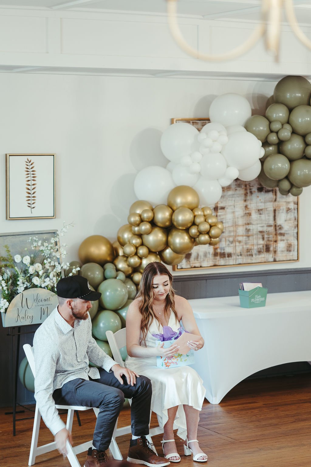 A couple sitting at a table in a decorated indoor venue with balloon arrangements in gold, white, and taupe, and a floral sign that reads "Welcome Kaley M".
