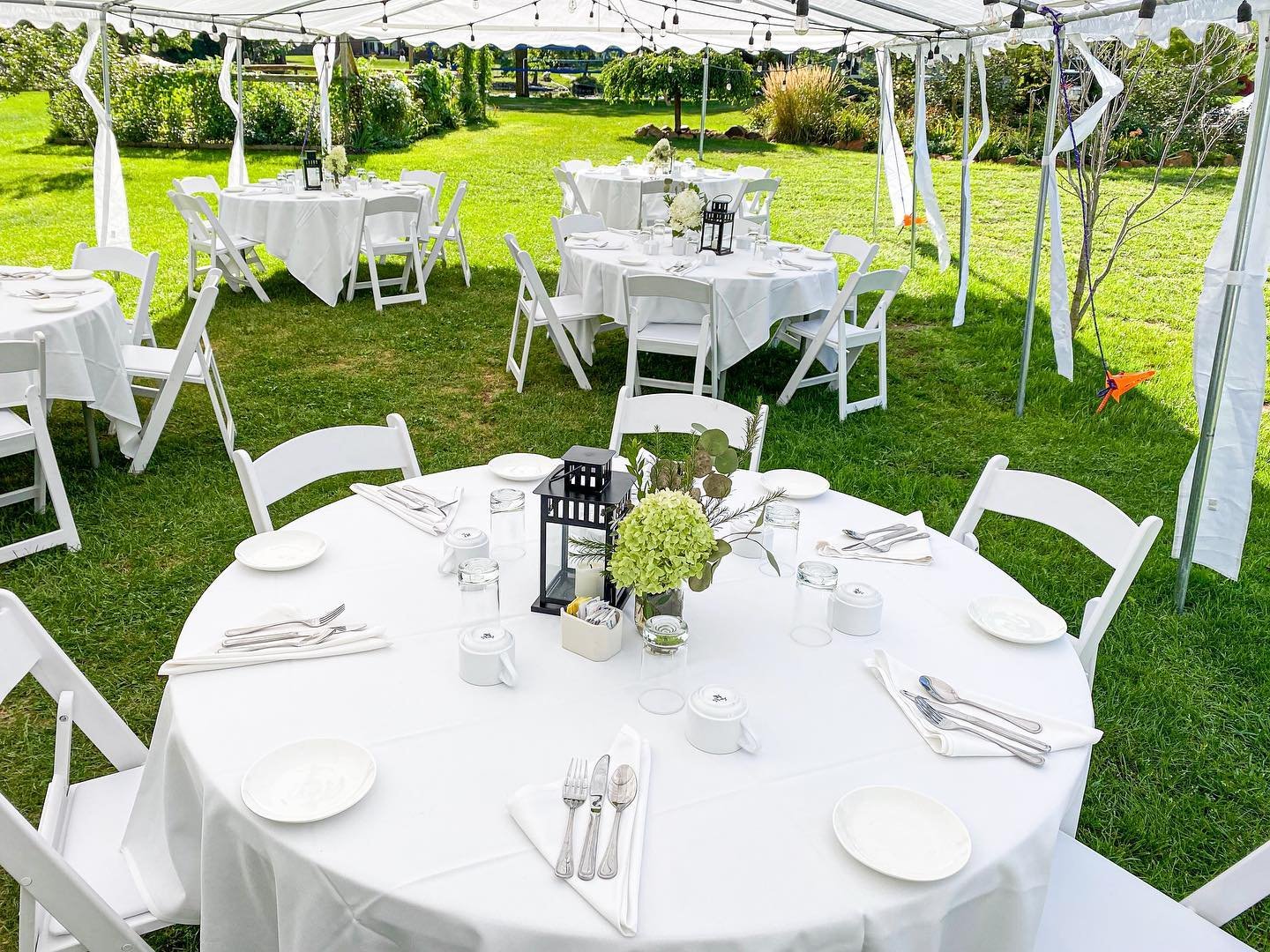 Outdoor event setup with round tables covered in white tablecloths, surrounded by white chairs, under a white canopy, with centerpieces, plates, glasses, and cutlery on the tables, on a grassy lawn with trees and shrubs in the background.
