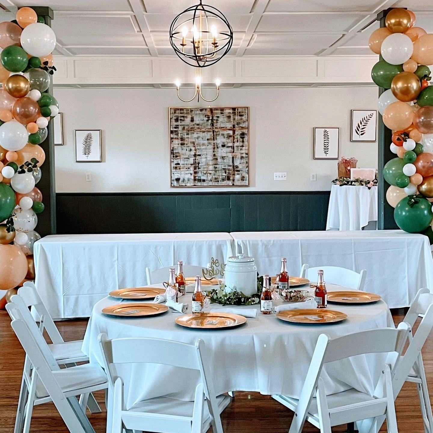 Decorated party room with a balloon arch in peach, white, green, and gold, white round tables with white chairs, gold plates, and bottles of soda, centerpiece with a white container and greenery, and artful wall decor.