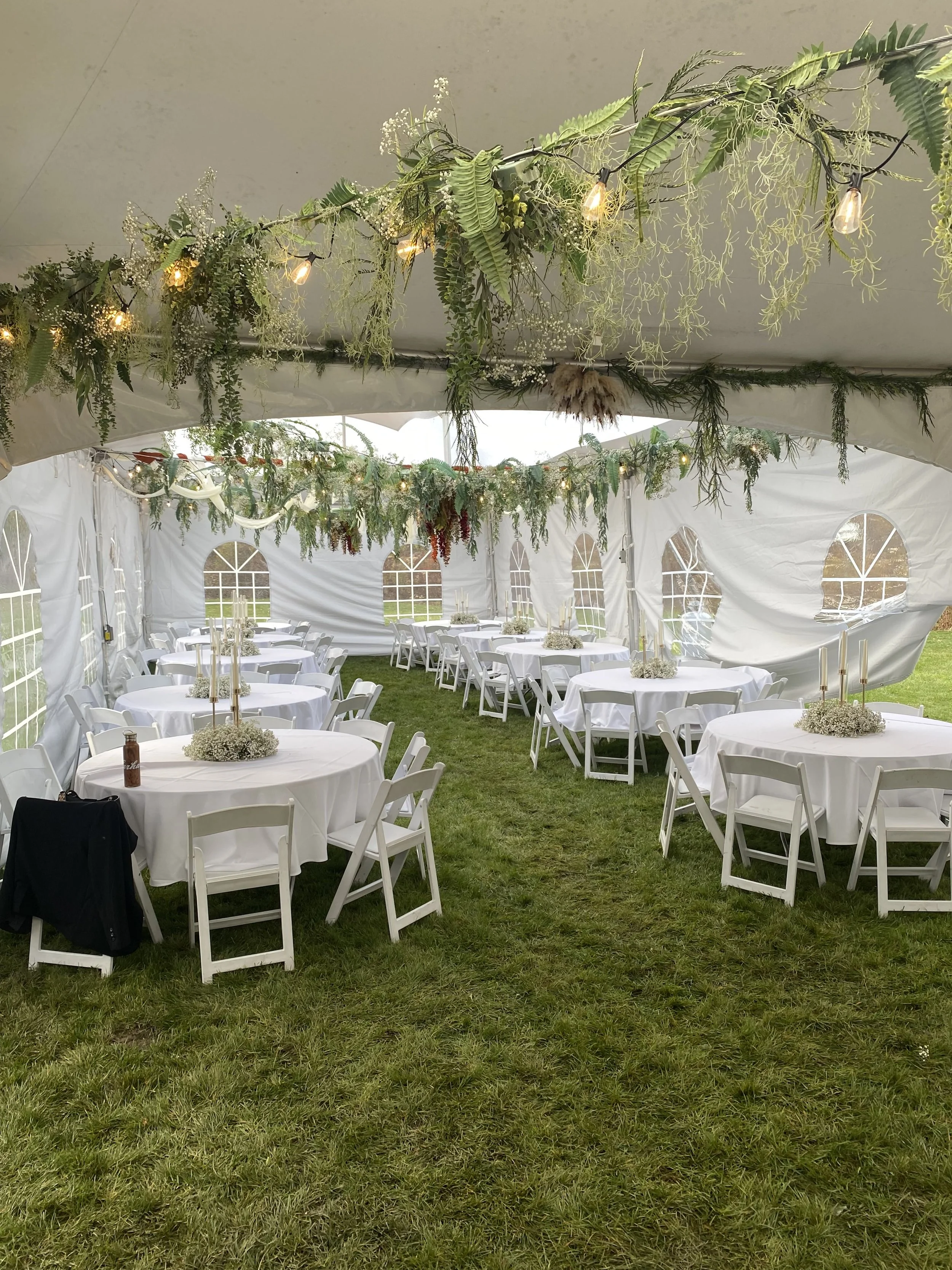 Inside a white event tent decorated with hanging green foliage and string lights, white round tables with white chairs are arranged on a grassy field for a celebration or wedding reception.