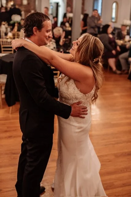 A bride and groom dancing at a wedding reception, with guests seated at tables in the background.
