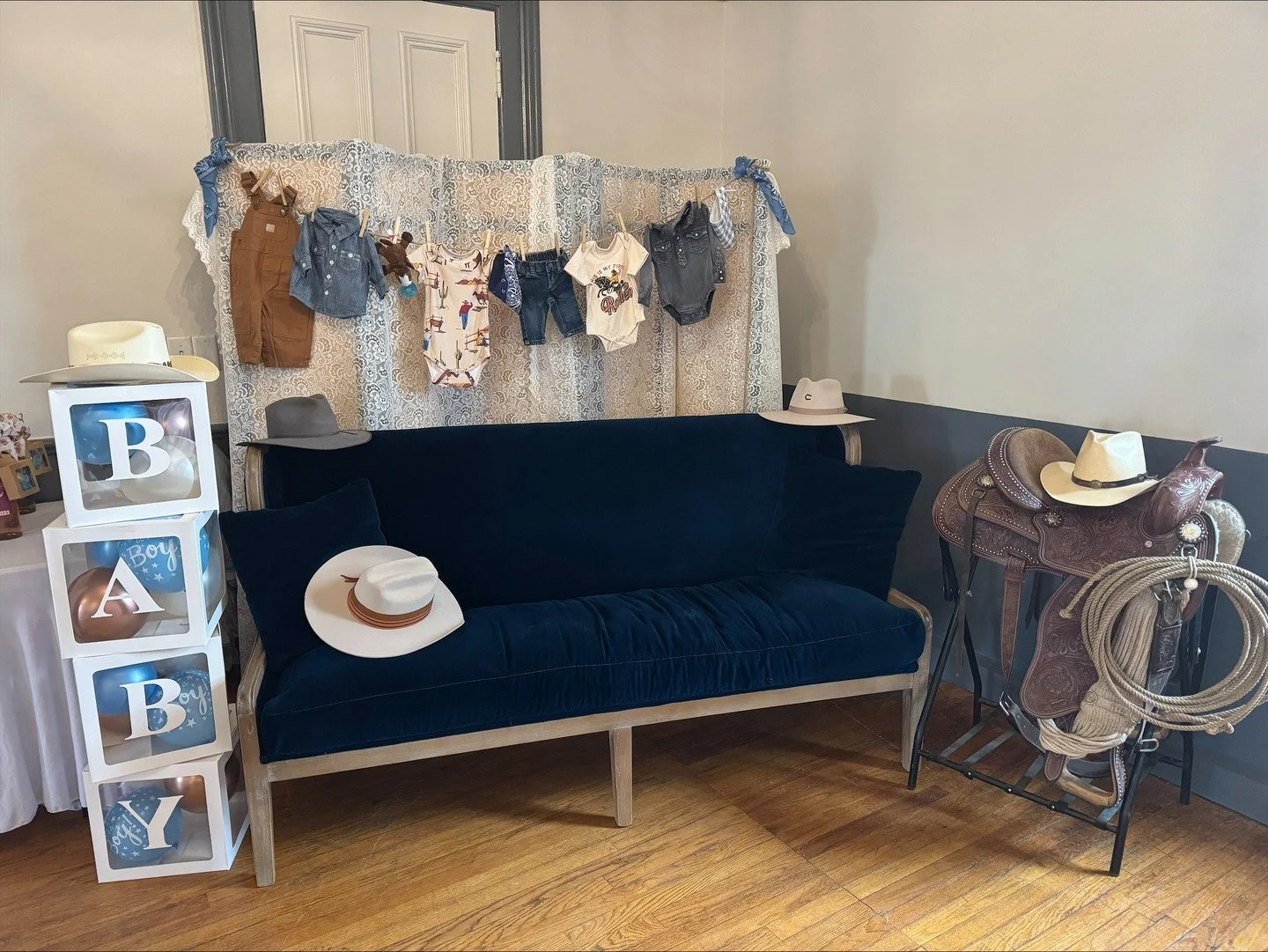 A black velvet couch with decorative pillows, surrounded by cowboy hats, a saddle, and a display of baby clothes hanging behind it.
