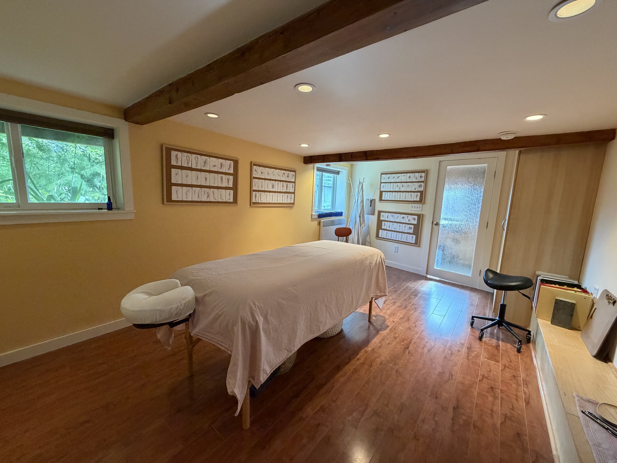 A massage table covered with a white sheet in a room with hardwood floors, yellow walls, window with greenery outside, and framed sketches on the wall. There is a small stool, a stool with a cloth draped over it, a coat rack, and a door with textured glass.